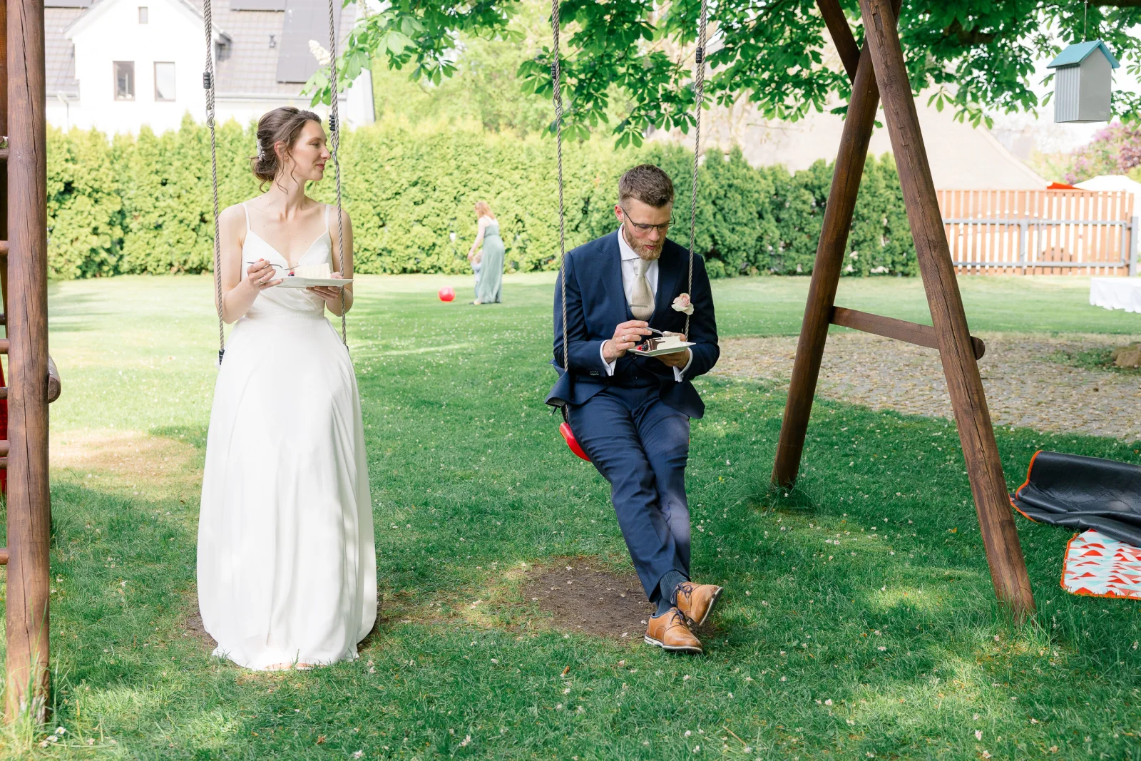 Ein Brautpaar isst im Garten Hochzeitstorte, der Bräutigam sitzt auf einer Schaukel.