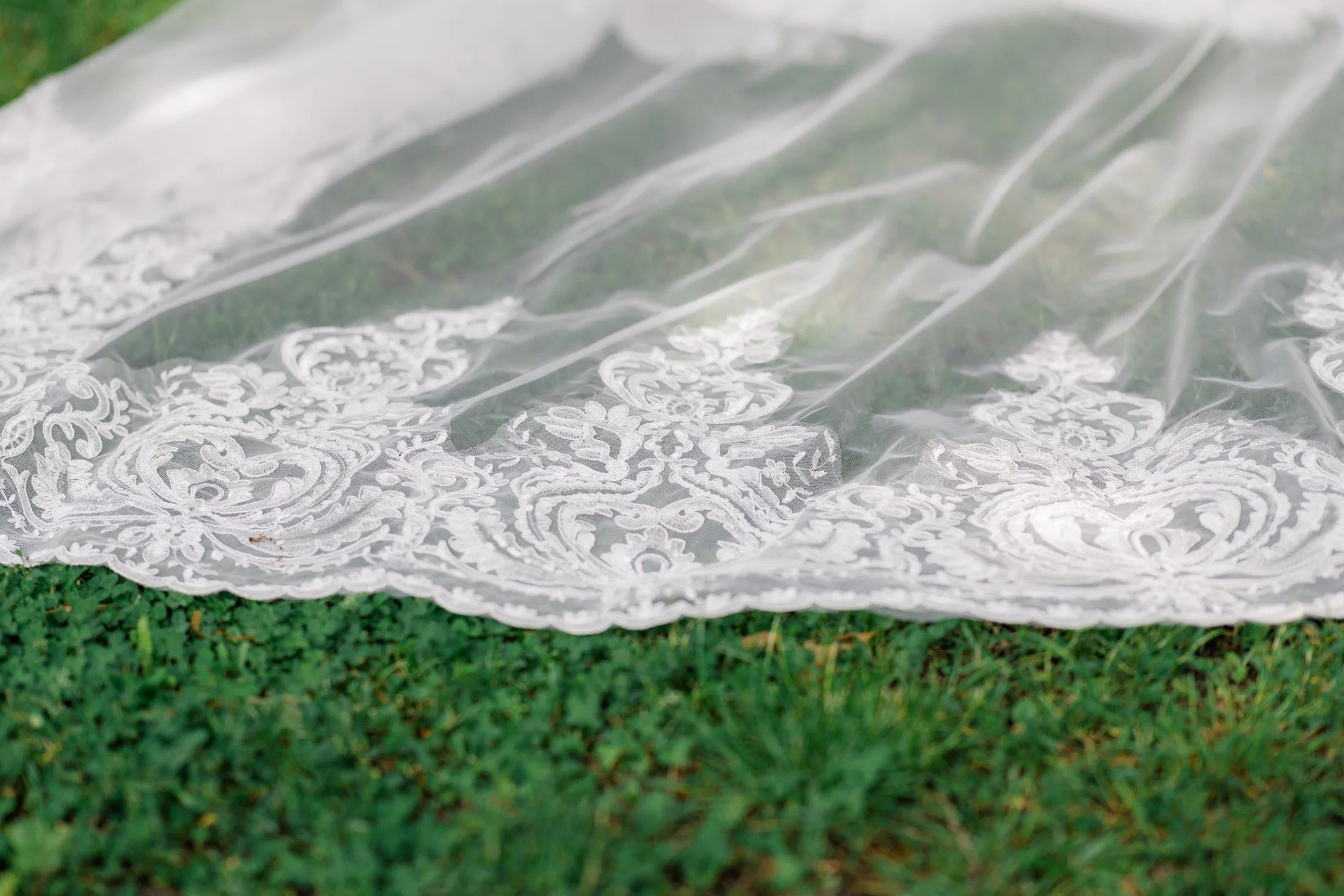 Die filigrane Spitze eines Brautkleids liegt auf grünem Rasen.